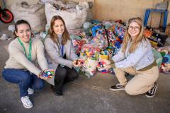 #ParaQuemNaoPodeVer Foto de três mulheres sorrindo, agachadas ou ajoelhadas próximas umas das outras. Elas estão segurando várias tampinhas plásticas coloridas nas mãos. Ao fundo, há vários sacos grandes cheios de tampinhas plásticas, sugerindo uma campanha de reciclagem ou coleta de tampas. O ambiente parece ser um galpão ou área de armazenamento. As três estão vestidas de forma casual e aparentam estar felizes com a atividade.