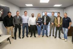 #ParaQuemNaoPodeVer Foto mostra um grupo de nove pessoas, todas em pé e organizadas em fila. O ambiente parece um escritório, com paredes claras, piso claro e janelas atrás do grupo. As pessoas estão vestidas de maneira casual a social, com camisas, jaquetas, calças jeans e algumas de camiseta. Há uma mulher no grupo enquanto o restante do grupo é composto por homens. Todos posam para a foto sorrindo. 