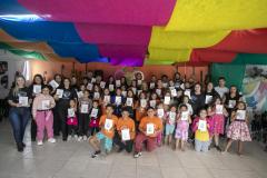 #praquemnaopodever foto mostra um grupo com 50 pessoas segurando um livro com capa branca posando para a foto. Estão em uma sala com parede laranja ao fundo e panos coloridos no teto. Ao fundo, uma um desenho de rosa com o nome 5C ao centro.