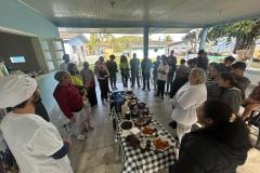 #praquemnaopodever foto mostra um grupo de pessoas em círculo em volta de uma mesa com toalha xadrez e repleta de comida. A maioria olha para uma pessoa que fala no outro lado da foto.