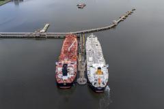#ParaQuemNaoPodeVer Foto aérea de dois navios-tanque atracados lado a lado em um píer no mar. O navio da esquerda é vermelho e o da direita é branco com detalhes em amarelo. Ambos estão alinhados paralelamente e conectados ao píer, que se estende da margem até o local onde eles estão. Próximo ao píer, à direita, há um pequeno barco navegando.