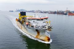 #ParaQuemNaoPodeVer Foto de um navio grande navegando em um corpo d'água, provavelmente próximo a um porto. O navio está indo em direção à direita da imagem e deixando um rastro na água. O navio é branco com detalhes amarelos e laranja, e parece equipado para operações industriais ou de transporte pesado, com guindastes e equipamentos grandes na parte traseira. Ao fundo, vê-se uma área portuária com guindastes, navios atracados e estruturas industriais próximas à água.