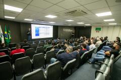 #ParaQuemNaoPodeVer Auditório com diversas pessoas sentadas em cadeiras assistindo a uma apresentação. Na frente, um homem está em pé próximo a um telão, onde uma apresentação de slides é projetada. Atrás dele, há bandeiras do Brasil e de outro estado ou instituição. O ambiente tem iluminação de teto forte, ar-condicionado e paredes claras. As pessoas parecem atentas à apresentação. A tela mostra informações em português, porém está levemente desfocada na imagem.