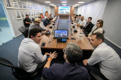 #praquemnaopodever foto mostra um homem sentado de costas para a camera, na ponta de uma mesa em formato de u e rodeado de pessoas sentadas com roupas formais olhando para ele.