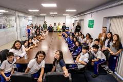 #ParaQuemNaoPodeVer Foto mostrando um grupo grande de pessoas sentadas ao redor de uma mesa de reunião em uma sala fechada. A maioria aparenta ser crianças e adolescentes, algumas usando uniformes escolares de calça azul e camiseta branca, enquanto adultos estão espalhados entre eles, talvez professores ou responsáveis. Todos olham para a câmera e sorriem, parecendo participarem de algum evento escolar ou visita guiada. No fundo, há quadros na parede e uma porta de vidro; do lado direito, há avisos e um car