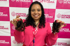 #praquemnaopodever foto mostra mulher negra de camisa rosa segurando uma mecha de cabelo de cada lado e sorrindo em frente de uma parede com os dizeres Portos do Paraná em rosa.