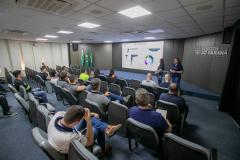 #ParaQuemNaoPodeVer Sala de apresentação com várias pessoas sentadas em cadeiras, assistindo a uma palestra. Na frente, há um telão exibindo gráficos e textos, além de duas pessoas apresentando. No fundo, há bandeiras do Brasil e do Paraná, e na parede está escrito "Portos do Paraná". O ambiente é bem iluminado, com teto branco e piso azul. A atenção das pessoas está voltada para o telão e os palestrantes.