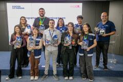 #ParaQuemNaoPodeVer Foto de um grupo de dez pessoas em pé, posando para a câmera em um ambiente interno, possivelmente em um evento ou cerimônia. Todas parecem estar sorrindo. Algumas seguram troféus e brindes, sugerindo premiação ou reconhecimento. O fundo tem um painel branco com texto, onde é possível ler “PORTOS DO PARANÁ”, “programa”, “digitalização”, e “HACK3R”. A maioria das pessoas usa crachás e roupas casuais ou uniformes. 