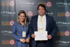 #ParaQuemNaoPodeVer Foto de um homem e uma mulher sorrindo, posando juntos para a câmera. Eles estão em frente a um painel de fundo decorado com os logos do "Programa Na Mão Certa" e da "Childhood Brasil", ambos destinados à proteção da infância. O homem está com um paletó azul, camisa branca e gravata amarela, segurando um certificado ou diploma. A mulher está usando uma roupa social azul claro, com crachá pendurado no pescoço. Ambos parecem estar participando de um evento formal ou solenidade de premiação