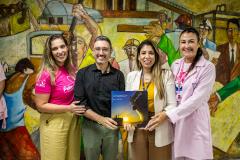 #praquemnaopodever foto mostra quatro pessoas lado a lado, em frente a um mural de trabalhadores, segurando um livro de capa azul e amarela escrito "Porto de Paranáguá - 90 anos"