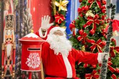 #praquemnaopodever foto mostra papai noel com barba branca comprida, lulas brancas e roupa vermelha com listra branca no meio sentado em frente a um painel com imagens natalinas. Papai Noel acena com a mão direita para a imagem, ao lado de uma arvore de natal cheia de adereços vermelhos.