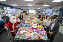 #praquemnaopodever a foto mostra uma mesa de madeira comprida e com quase mil brinquedos em cima de todas as cores e formatos. Nas lateriais, dezenas de pessoas estão a postos sorrindo e posando para o fotógrafo dentro de uma sala de reunião com tapete cinza e janelas grandes