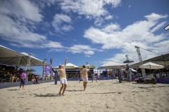 #praquemnaopodever imagem na praia, dentro da quadra de areia, a dupla de homens de bermuda branca e camisa amarela estão a frente de costas pra foto. Do outro lado está a outra dupla pronta pra receber a bola. O céu está azul com algumas nuvens. Ao fundo e dos lados as arquibancadas estão lotadas