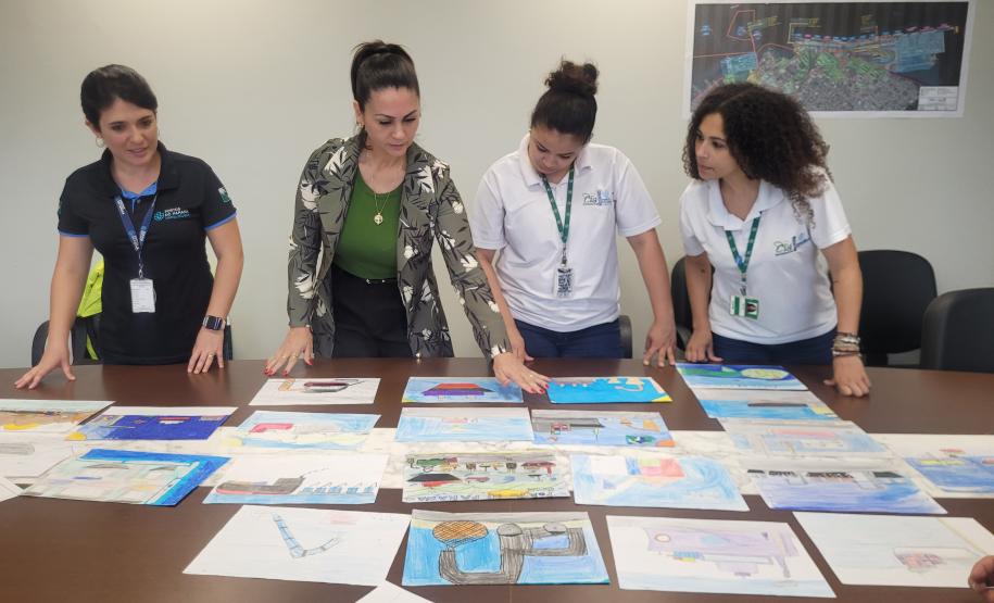 Quatro mulheres lado a lado olham para uma mesa com várias folhas cheias de desenhos de portos, navios e animais. Uma delas aponta para uma folha e as outras acompanham na direção do dedo. Todas estão concentradas, escolhendo os melhores desenhos e frases.