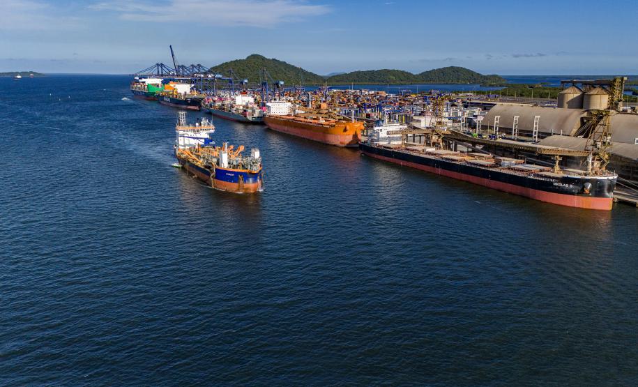 #paraquemnaove Imagem mostra grande parte do mar azul à esquerda, e à direita está o Porto de Paranaguá com navios atracados por todo o cais. No mar, segue navegando um navio vermelho de com faixa azul. O céu está aberto, com poucas nuvens.