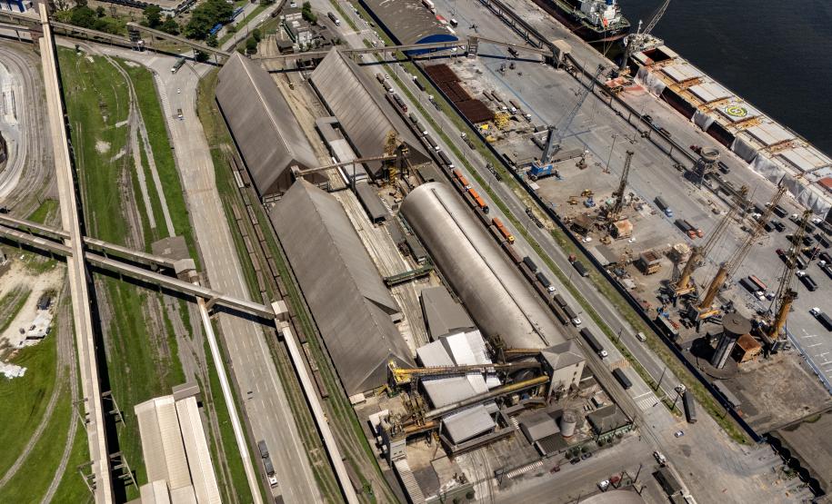 #ParaQuemNaoPodeVer Na imagem, é possível ver várias estruturas grandes, como armazéns ou silos, além de guindastes e outras máquinas pesadas. Há também uma área de cais onde estão atracados navios ou embarcações.