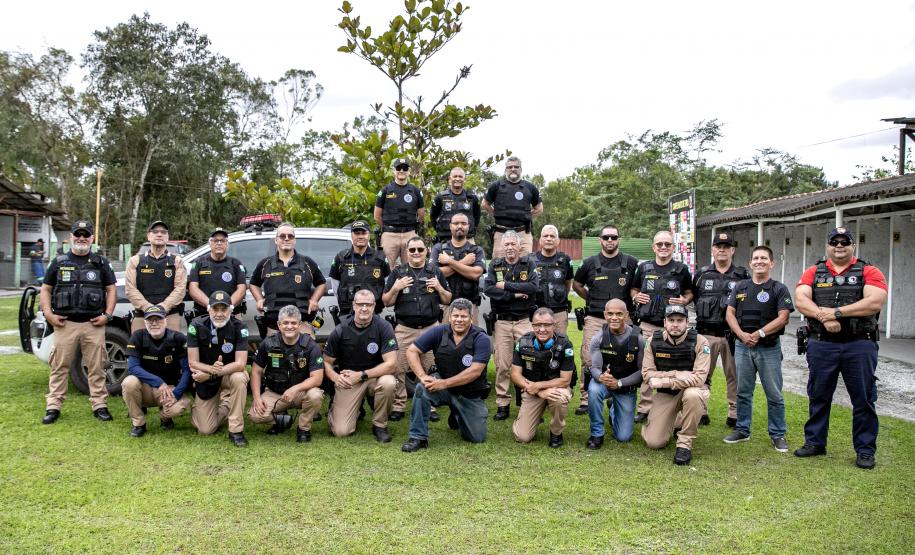 Dezenas de guardas portuárias estão fardados e posando para a foto lado a lado em um gramado. A maioria veste uma calça creme com camisa preta e colete, com os braços cruzados à frente.