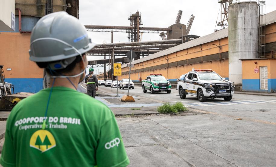 #ParaQuemNaoPodeVer A imagem mostra um homem em primeiro plano, visto de costas, usando um capacete de segurança cinza e uma camisa verde. Ao fundo, há uma estrutura industrial com um prédio laranja e azul e duas viaturas. A primeira, traz na carroceria a imagem de Nossa Senhora do Rocio. Atrás das viaturas, seguem outros veículos em fila, acompanham o cortejo pelas instalações do Porto de Paranaguá.