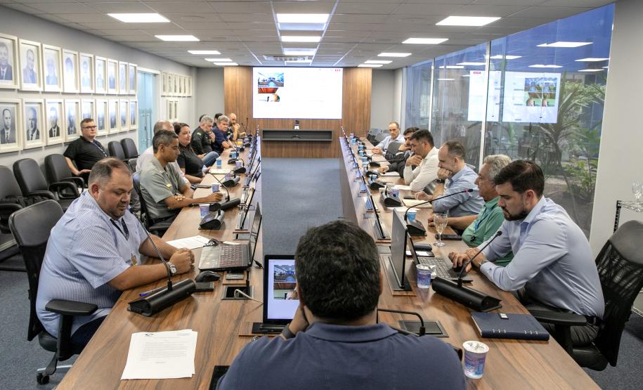 #ParaQuemNaoPodever Na foto, há várias pessoas sentadas ao redor de uma longa mesa de conferência. As pessoas estão usando roupas formais e parecem estar participando de uma reunião ou discussão. Há microfones na mesa em frente a cada pessoa, e algumas delas estão usando laptops ou tablets. A sala tem uma parede de vidro ao fundo, através da qual é possível ver plantas. Na parede oposta, há quadros com retratos de pessoas. A iluminação é feita por lâmpadas embutidas no teto.