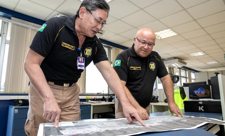 Na imagem, dois guardas portuários estão lado a lado olhando para um mapa sobre uma mesa azul. O da esquerda aponta para a imagem enquanto o da direita observa a imagem.