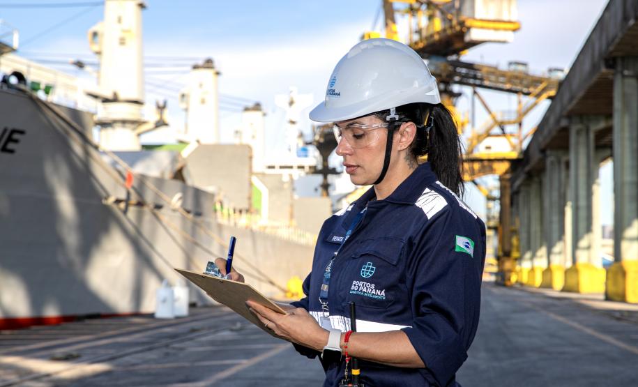 Portuária de uniforme azul de manga com o logo estampado "portos do parana" escreve em uma prancheta com a mão esquerda, enquanto um navio cinza ao fundo é carregado. Ela está de capacete branco e usa epis completos. #paraquemnaopodever