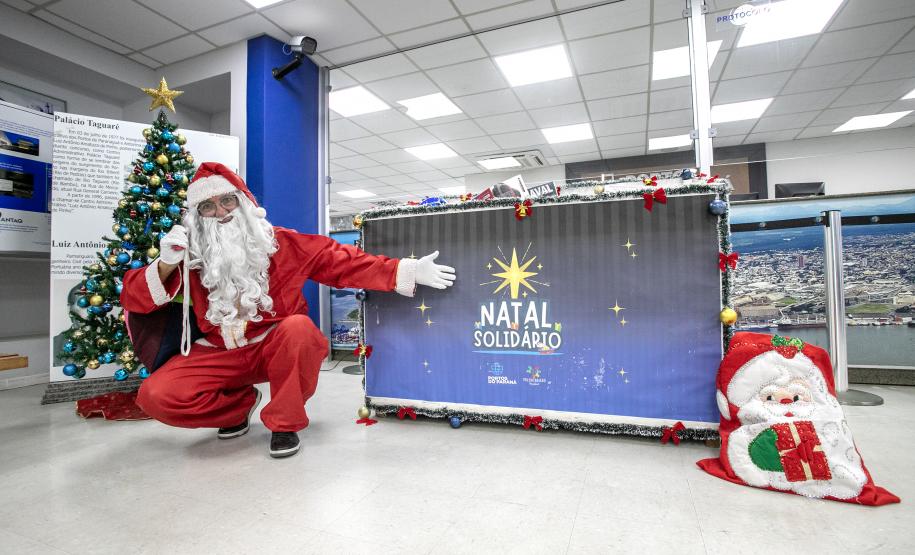 #ParaQuemNaoPodeVer A imagem mostra um homem vestido de Papai Noel agachado ao lado de uma árvore de Natal decorada com bolas azuis e douradas e uma estrela dourada no topo. Ele está usando um traje vermelho com detalhes brancos, incluindo um gorro e uma longa barba branca. O homem está fazendo um gesto de "paz e amor" com a mão direita. Ao lado dele, há um painel azul com a inscrição "Natal Solidário" em letras brancas e uma estrela amarela acima do texto. Na frente do painel, há um tapete ou almofada com