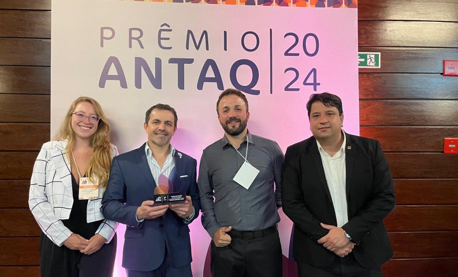 Quatro pessoas estão lado a lado, em frente a uma parede de madeira com um banner escrito "prêmio antaq 2024". São três homens e uma mulher com roupas formais, sorrindo e um deles segura um prêmio nas mãos. #praquemnaove