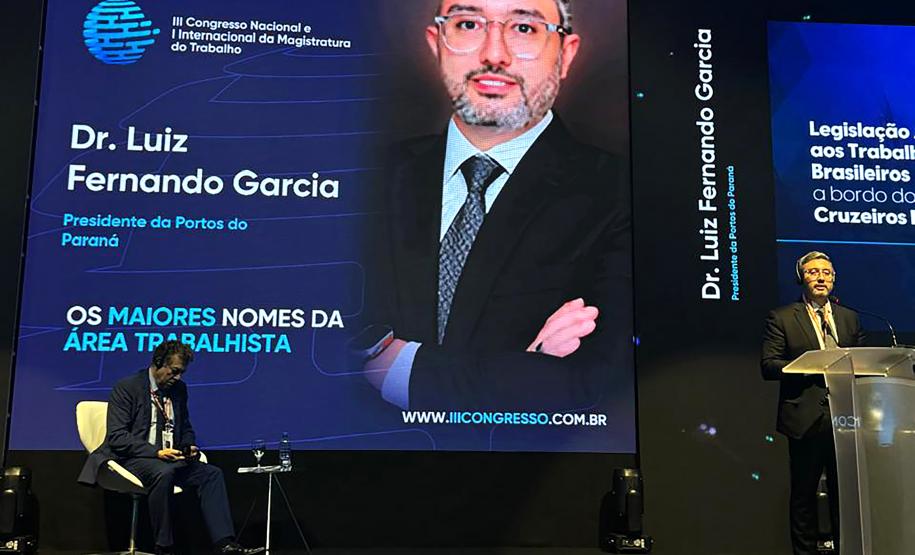 #ParaQuemNaoPodever A imagem mostra uma apresentação ou conferência. No centro da tela, há um slide com fundo azul escuro e texto em branco e azul claro. O texto diz "Dr. Luiz Fernando Garcia" e "Presidente da Portos do Paraná". Abaixo, está escrito "OS MAIORES NOMES DA ÁREA TRABALHISTA". No canto inferior direito do slide, há um logotipo circular azul com linhas horizontais.  Na parte inferior da tela, há uma foto de um homem de terno e gravata, que parece ser o Dr. Luiz Fe