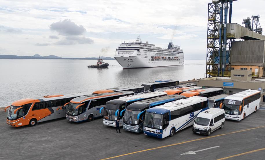 #praquemnaopodever 7 ônibus e uma van estão a frente da foto, no cais do Porto de Paranaguá, aguardando a chegada, atrás, do imponente navio de cruzeiros MSC Armonia.