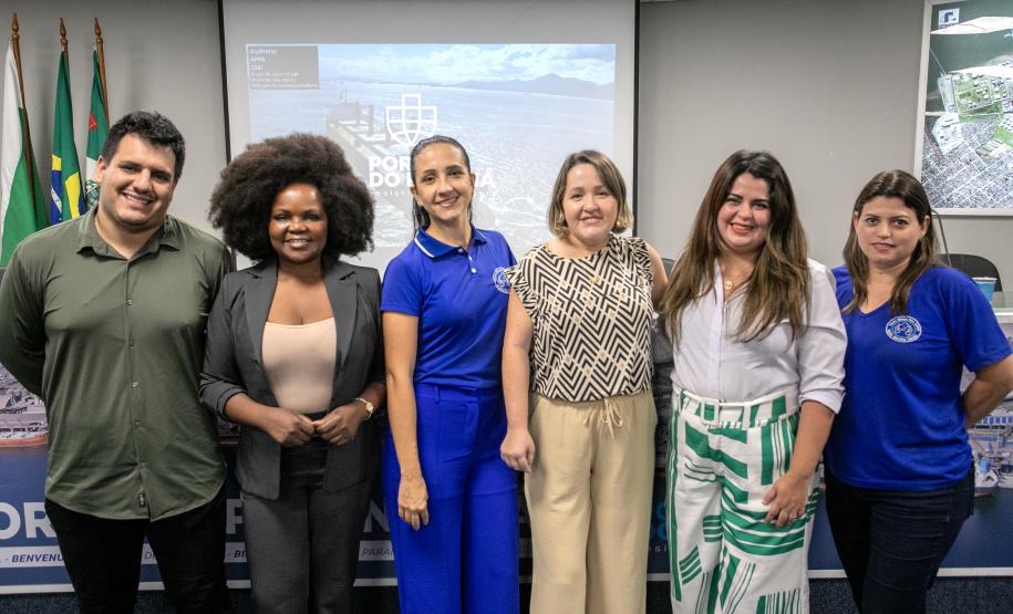 #ParaQuemNaoPodeVer A imagem mostra seis pessoas posando para uma foto em um ambiente interno. Há um homem de camisa verde e duas mulheres, uma com cabelo afro e outra com cabelo preso. Na fileira da frente, há três mulheres, uma usando uma camisa azul, outra com uma blusa estampada e a terceira com uma blusa branca e saia com listras verdes. Ao fundo, há bandeiras e um painel que parece ter um mapa ou uma imagem de cidade.