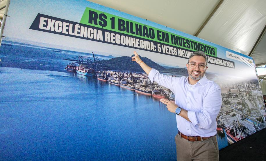 #paraquemnaopodever Homem de camisa banca, calça marrom e cinto marrom aponta para outdoor atrás com frase "R$ 1 Bilhão em investimentos"
