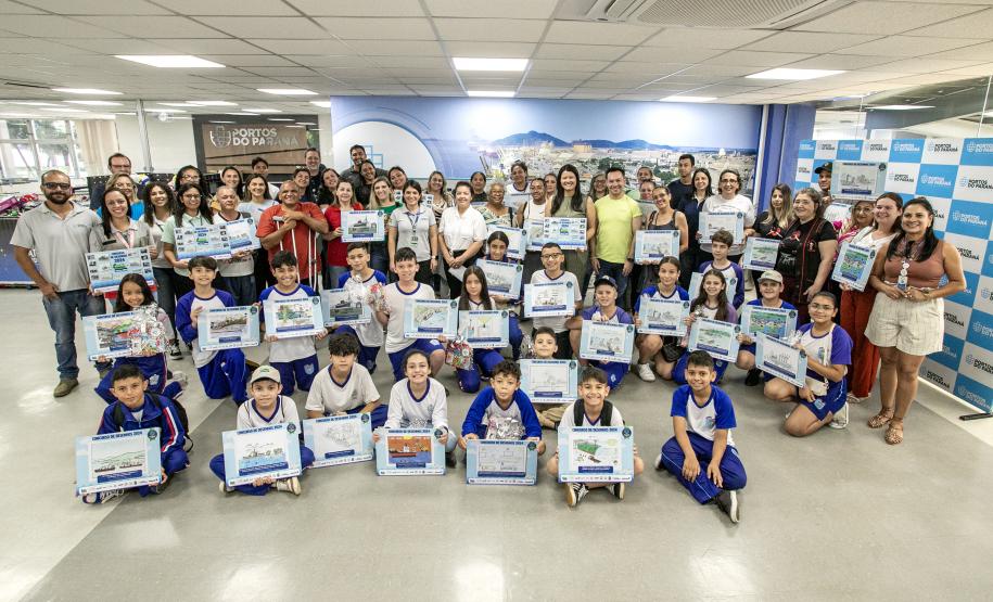 #praquemnaopodever Na recepção da Portos do Paraná, dezenas de crianças estão posicionadas segurando uma placa em homenagem ao prêmio do Concurso de Desenho do Porto Escola. Professores, pais, representantes dos municípios e da Portos do Paraná também estão na foto.