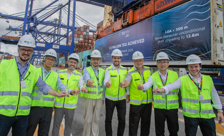 #paraquemnaopodever nove homens de coletes verdes e capacetes brancos seguram taças de champagne com os braços esticados para a frente. Ao fundo, guindastes azuis de mais de 50 metros de altura movimentam contêineres e à direita um outdoor em um contêiner com os dizeres "1,5 million TEU achieved"