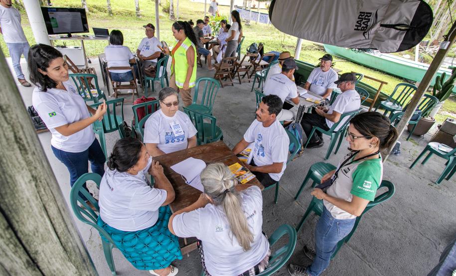 #praquemnaopodever várias pessoas sentadas em cadeiras de plástico verdes separadas em grupos gesticulam sobre projetos socioambientais. Todos vestem camisas brancas e conversam sérios enquanto outras pessoas em pe passam pelas pesas. Um prancha está presa ao teto e ao fundo é possível vera grama da Ilha do Teixeira
