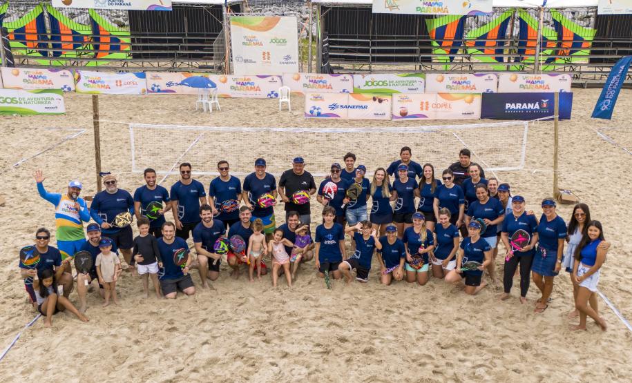 #ParaQuemNaoPodeVer A imagem mostra um grupo grande de pessoas posando para uma foto em uma quadra de areia, possivelmente de vôlei de praia. Todos estão vestindo camisetas azuis e alguns seguram bolas coloridas. Ao fundo, há uma estrutura com várias faixas e banners, que parecem ser de patrocinadores ou eventos, com logotipos e textos.