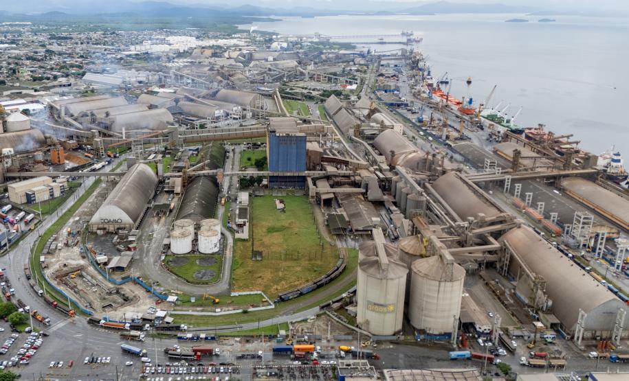 #ParaQuemNaoPodeVer A imagem mostra uma foto aérea de uma área portuária. Na imagem, é possível ver várias estruturas grandes, como tanques cilíndricos e prédios industriais, além de áreas verdes ao redor. Há também várias estradas e veículos visíveis.