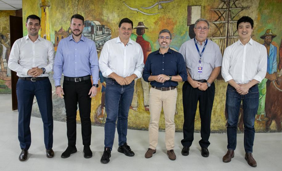 #ParaQuemNaoPodeVer A imagem mostra uma foto de seis homens em pé, posando para a câmera. Eles estão em duas fileiras, três na frente e três atrás. Todos estão vestidos de forma formal, com camisas sociais e calças. O fundo da foto é uma pintura mural colorida que parece representar uma cena cultural ou histórica.