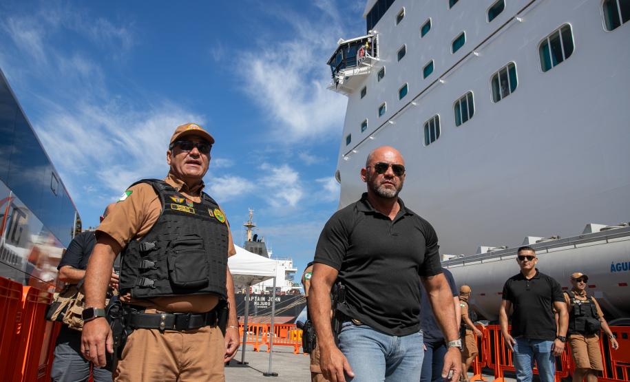 #ParaQuemNaoPodeVer A imagem mostra dois homens em destaque. O homem à esquerda está usando um uniforme marrom com um colete preto e um boné, além de óculos escuros. Ele parece ser um oficial de segurança. O homem à direita está vestindo uma camisa preta e também usa óculos escuros. Ao fundo, há um grande navio de cruzeiro branco, com várias janelas visíveis, e o céu está azul com algumas nuvens.