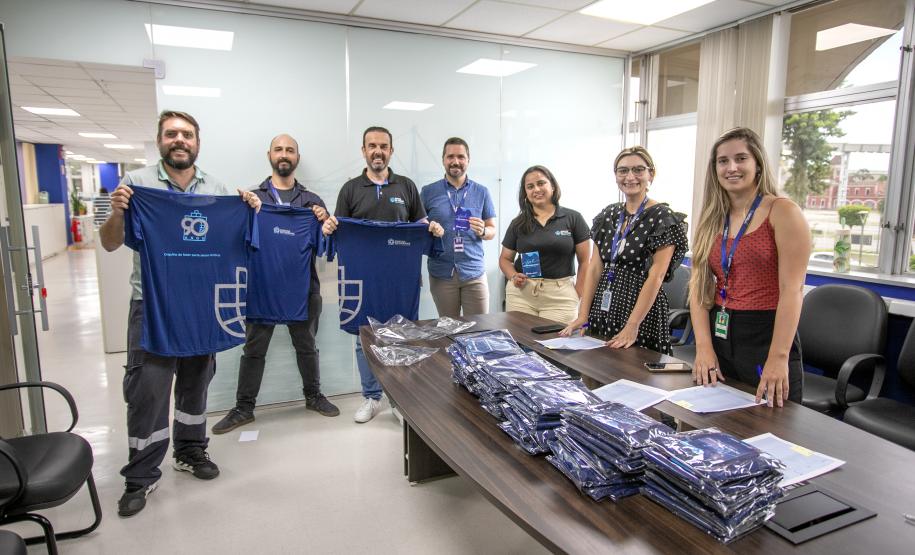 #ParaQuemNaoPodeVer A imagem mostra um grupo de pessoas em um ambiente de escritório. Na parte superior da imagem, há quatro homens em pé, segurando camisetas azuis com um logotipo branco. Eles estão sorrindo e parecem estar participando de algum tipo de evento ou celebração. Na parte inferior da imagem, há três mulheres e um homem em pé atrás de uma mesa, que está coberta com várias pilhas de camisetas azuis dobradas. As mulheres estão sorrindo e também parecem estar participando do mesmo evento.