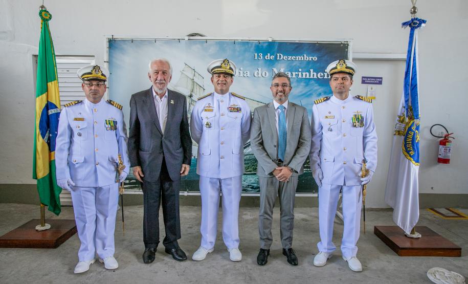 #ParaQuemNaoPodeVer A imagem mostra cinco homens posando para uma foto em frente a uma bandeira do Brasil e uma bandeira da Marinha do Brasil. Três deles estão vestidos com uniformes brancos de oficiais da Marinha, com chapéus brancos e insígnias no peito. Os outros dois homens estão usando ternos. Ao fundo, há um banner azul com o texto "13 de Dezembro" e "Dia do Marinheiro". A foto parece ter sido tirada em um ambiente interno, com uma parede branca ao fundo.