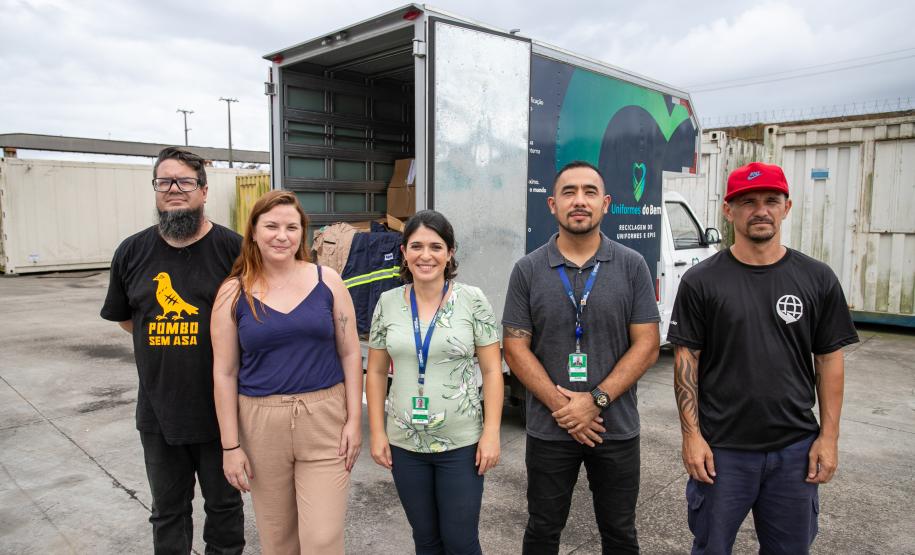 #praquemnaopodever cinco pessoas lado a lado posam para a foto em frente a um caminhão azul e verde que está com as portas da caçamba aberta e cheia de caixas com roupas.