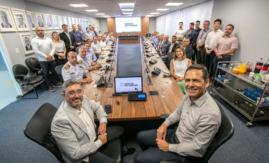 #praquemnaopodever em torno de uma mesa de madeira em formato de u, muitos homens e poucas mulheres posam para a lente da câmera. Eles estão na sala da Portos do Paraná. Com roupas formais, alguns estão sentados, outros em pé, ao fundo aparece o logo Portos do Paraná no telão da exibição.