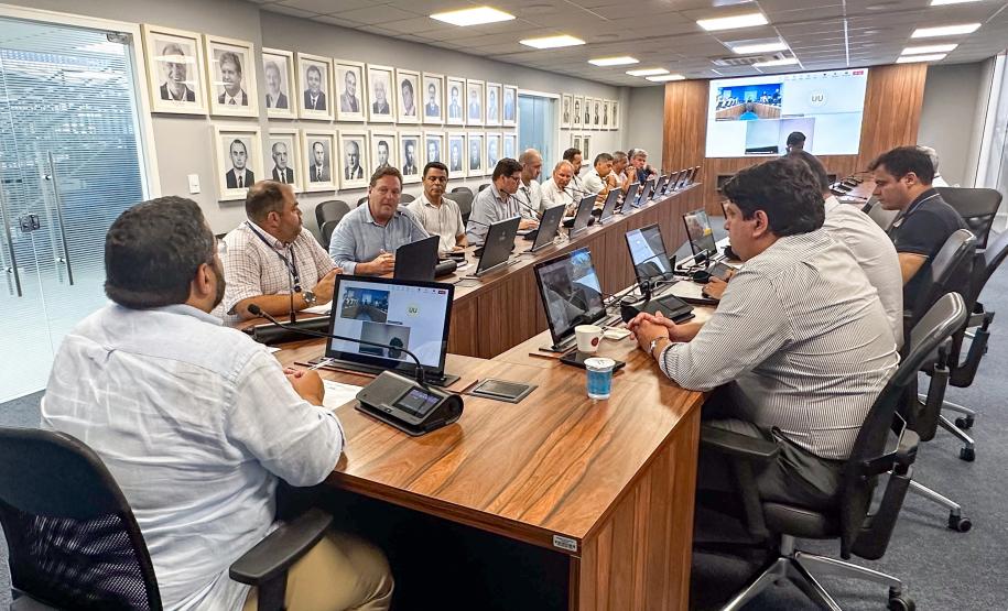 #ParaQuemNaoPodeVer A imagem mostra uma foto de uma reunião em uma sala de conferências. Há várias pessoas sentadas ao redor de uma mesa de madeira, todas olhando para a frente, onde parece haver uma apresentação ou videoconferência em andamento. Na parede ao fundo, há várias fotos emolduradas. As pessoas estão usando roupas formais, como camisas sociais. Na mesa, há laptops, garrafas de água e outros materiais de escritório. A sala é bem iluminada, com luzes no teto.