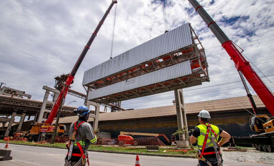 #ParaQuemNaoPodeVer A imagem mostra uma fotografia de um local de construção. Na foto, há dois trabalhadores usando equipamentos de segurança, como capacetes e coletes refletivos. Eles estão observando uma grande estrutura metálica sendo levantada por um guindaste vermelho. Ao fundo, há uma construção industrial com várias vigas e estruturas de aço. O céu está nublado.
