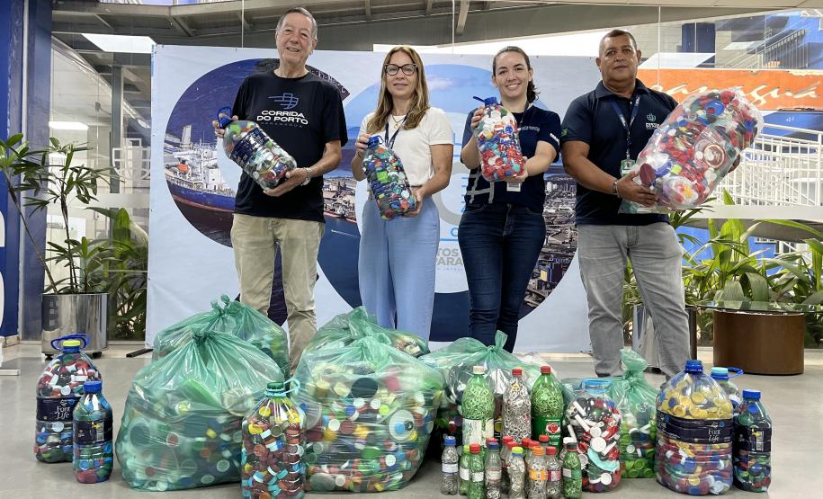#ParaQuemNaoPodeVer Quatro pessoas estão posando para uma foto em um ambiente interno. Elas estão segurando garrafas grandes e sacos cheios de tampinhas plásticas coloridas. No chão, há mais sacos e garrafas cheias das mesmas tampinhas. As pessoas estão sorrindo e parecem estar participando de uma campanha de coleta de tampinhas. Ao fundo, há um banner com uma imagem de um navio e algumas plantas decorativas. As pessoas estão vestindo camisetas pretas com um logotipo branco.