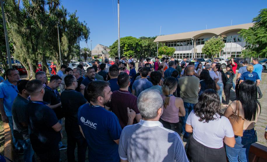 #ParaQuemNaoPodeVer A foto mostra um grupo grande de pessoas reunidas ao ar livre, em um local que parece ser uma área urbana com árvores e um prédio ao fundo. As pessoas estão em fila ou aglomeradas, e o céu está claro e azul.