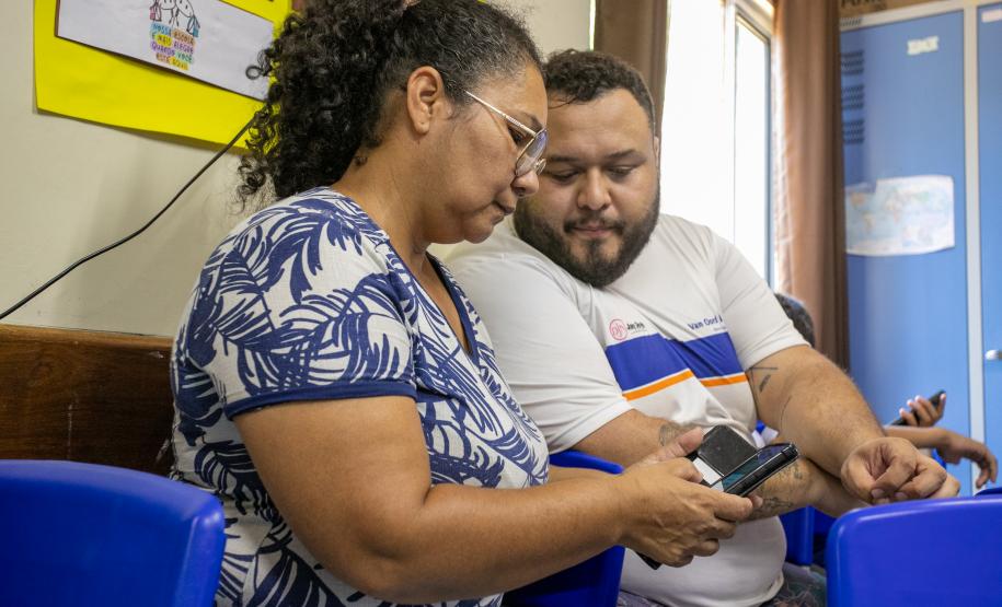 #praquemnaopodever uma mulher negra de blusa branca com plantas azuis mexe no celular ao lado de um rapaz de camisa branca com listras azul e laranja. Os dois estão sentados dentro de uma sala de aula e olham para o aparelho, enquanto a senhora de meia idade e óculos digita no celular.
