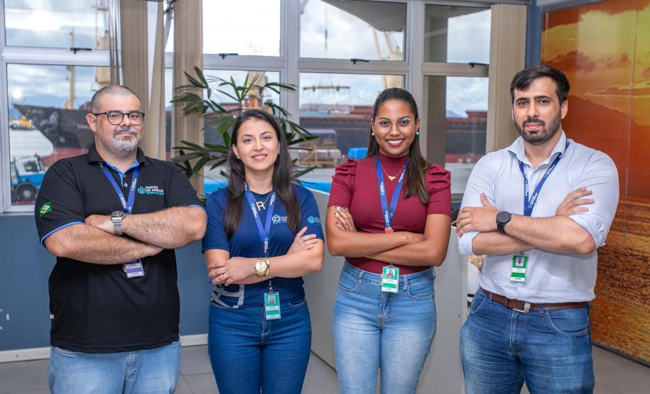 #ParaQuemNaoPodeVer Imagem exibe quatro pessoas posando juntas. As pessoas estão em pé, duas na frente e duas atrás, todas com os braços cruzados. A pessoa à esquerda, na parte de trás, é um homem usando uma camisa preta com detalhes em azul e jeans. Ele tem cabelo curto e barba. Ao lado dele, à direita, está uma mulher com cabelo escuro e liso, vestindo uma camisa azul. Na frente, à esquerda, está uma mulher com cabelo preso, usando uma blusa vermelha. À direita, na frente, está um homem com cabelo escuro