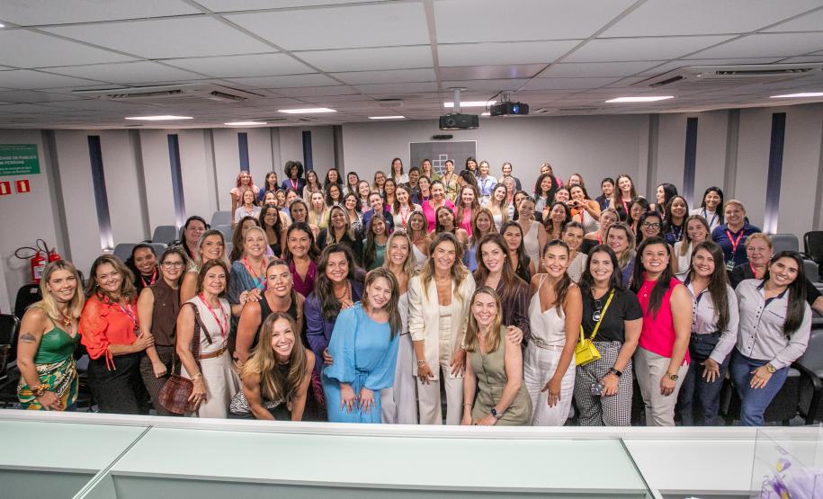 #ParaQuemNaoPodeVer Imagem mostra um grande grupo de pessoas, principalmente mulheres, posando juntas em um ambiente interno, possivelmente uma sala de conferências ou auditório. As pessoas estão sorrindo e parecem estar participando de um evento ou reunião. Algumas estão vestindo roupas coloridas e outras em tons mais neutros.