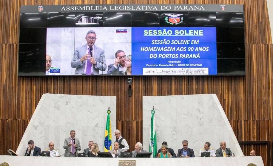 #praquemnaopodever 10 pessoas estão sentadas em um púlpito alto e com fachada branca. Em pé está um homem de terno cinza e gravata rosa, segurando um microfone na mão e discursando enquanto gesticula com a mão direita. Seu perfil aparece no telão ao fundo, ao lado de um texto com os dizeres "sessão solene em homenagem aos 90 anos do Portos Paraná"